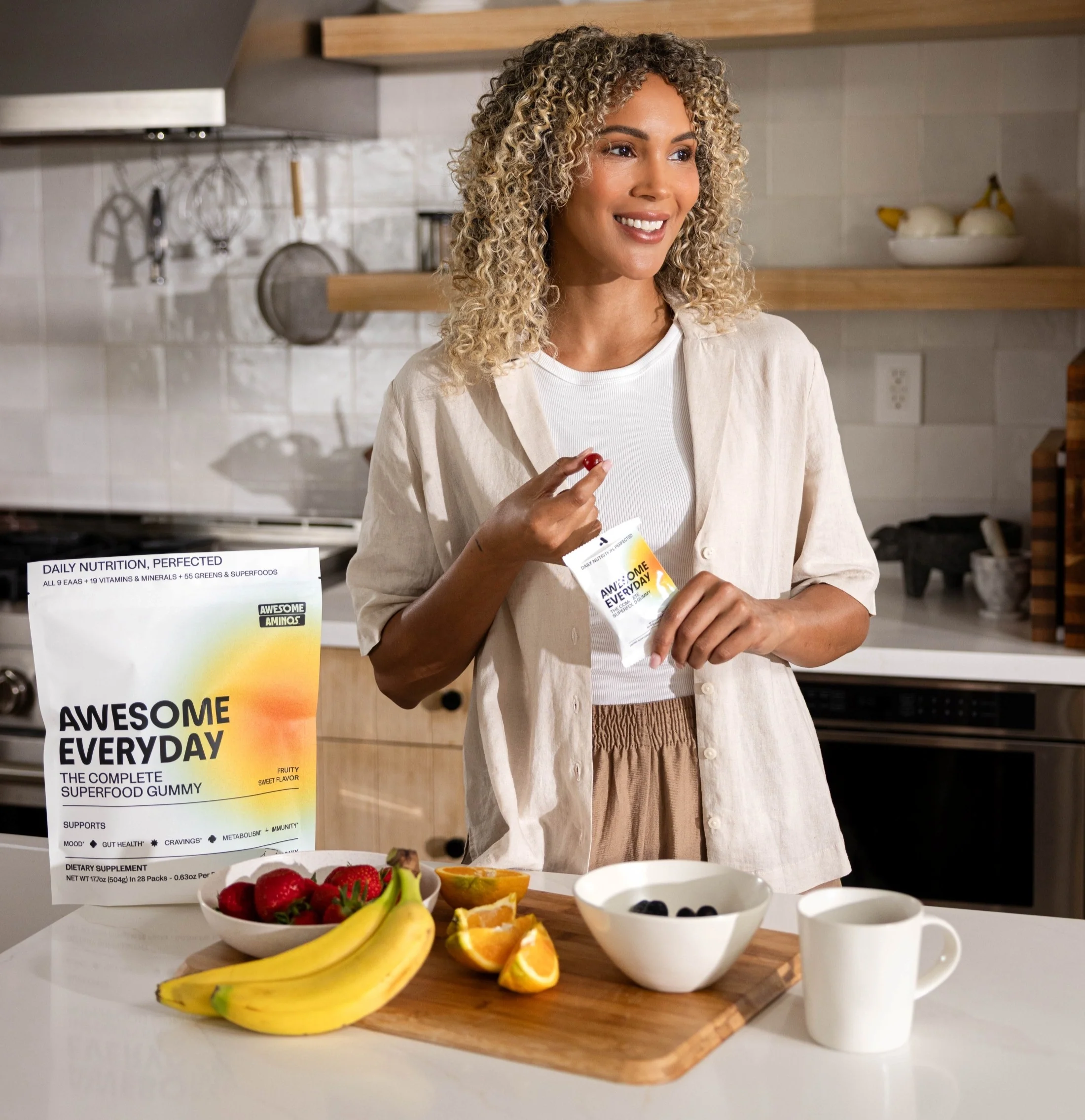Woman holding an Awesome Everyday™ gummy pouch beside fresh fruit in a kitchen.